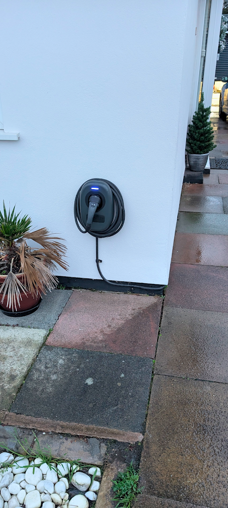 Black EV charger with coiled cable on a white wall beside a tiled path
